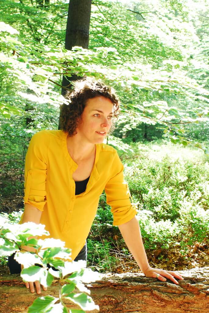 Een vrouw met een gele blouse aan die op een boomstam in het bos leunt en in de verte kijkt.